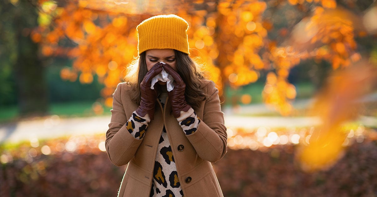 Lady sneezing outside around falling leaves.